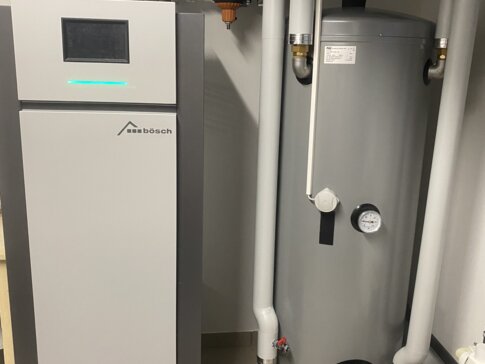 Sole/Wasser-Wärmepumpe KLIMT von bösch in einem Heizungsraum, neben einem großen grauen Warmwasserspeicher. Die Wärmepumpe ist mit einem digitalen Display ausgestattet und verbunden mit mehreren Rohrleitungen, Ventilen und Manometern zur Steuerung der Temperatur und Wasserzirkulation. | © Walter Bösch GmbH & Co KG