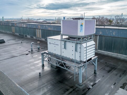 Luftaufnahme eines großen, auf einem Fabrikdach installierten Lüftungssystems. Die Anlage steht auf einer Metallstruktur und ist vorwiegend in Weiß gehalten, mit einem oberen Gitter in Silber. Das Dach ist nass, möglicherweise nach einem Regenschauer. Im Hintergrund ist eine städtische Umgebung mit entfernten Gebäuden und leicht bewölktem Himmel zu erkennen. | © INFRANORM®