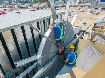 Zwei Arbeiter auf einem Dach arbeiten an der Montage eines großen, metallischen Lüftungssystems. Sie sind mit Sicherheitsausrüstung ausgestattet und konzentrieren sich auf ihre Aufgabe.