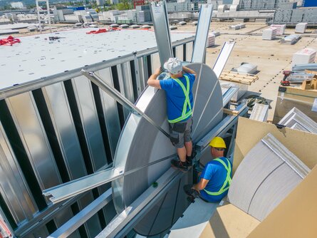 Zwei Arbeiter auf einem Dach arbeiten an der Montage eines großen, metallischen Lüftungssystems. Sie sind mit Sicherheitsausrüstung ausgestattet und konzentrieren sich auf ihre Aufgabe.