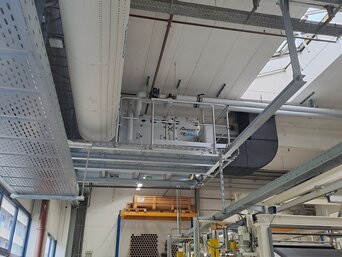 Blick auf eine industrielle Produktionshalle mit großen Rohrleitungen und Lüftungssystemen an der Decke. Unten befinden sich Maschinen und Regale mit Materialien. Im Hintergrund sind gelbe und weiße Maschinen zu sehen, die Teil des Produktionsprozesses sind. | © Infranorm, Wels (AT)