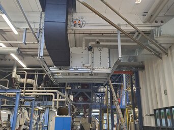 Industrielle Produktionshalle mit komplexen Rohrleitungen und einer großen, schwarzen Lüftungsanlage an der Decke. Unten arbeitet eine Person an einem Tisch mit einem Laptop, umgeben von Maschinen und technischen Geräten. Im Hintergrund sind Leitern und weitere technische Strukturen zu sehen. | © Infranorm, Wels (AT)