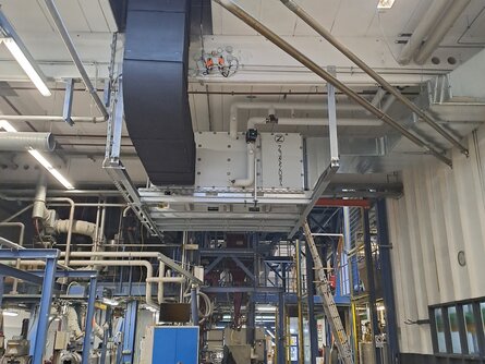 Industrielle Produktionshalle mit komplexen Rohrleitungen und einer großen, schwarzen Lüftungsanlage an der Decke. Unten arbeitet eine Person an einem Tisch mit einem Laptop, umgeben von Maschinen und technischen Geräten. Im Hintergrund sind Leitern und weitere technische Strukturen zu sehen. | © Infranorm, Wels (AT)