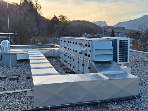 Ein Dach mit mehreren Klimaanlagen-Einheiten in verschiedenen Größen und Formen bei Sonnenuntergang. Die Anlagen sind auf einem mit Kies bedeckten Dach platziert, das sich vor einem Hintergrund mit bewaldeten Bergen erstreckt. Der Himmel ist leicht bewölkt und färbt sich orange durch die untergehende Sonne.
